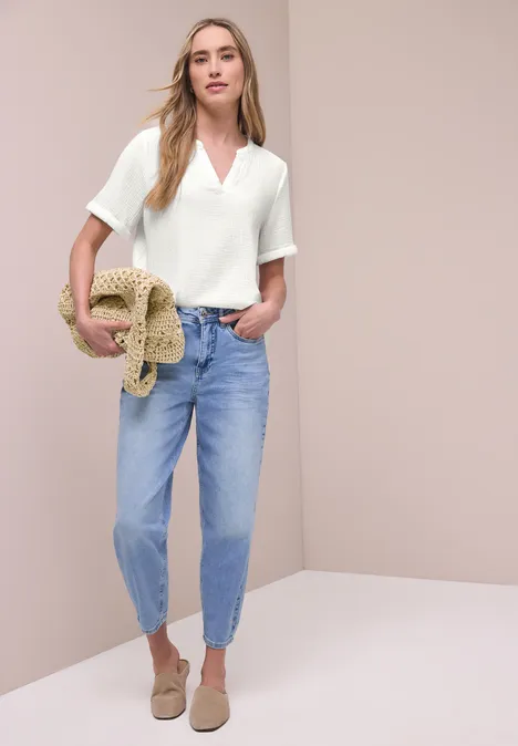 Frau mit weißem V-Shirt, hellblauen Jeans, hellbraunen Slippern, hält Strohhut und Clutch vor beigem Hintergrund.